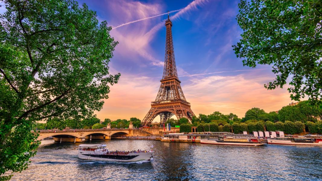 The Eiffel Tower in Paris, France, viewed across the Seine River with sightseeing boats on the water, trees along the riverbanks, and a colorful sunset sky in the background