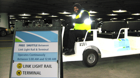 An operator smiles in the driver's seat of a shuttle behind a placard displaying shuttle ride hours