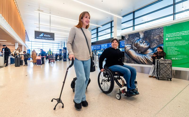 A traveler using a cane and traveler in a wheelchair walk through the terminal at SEA.