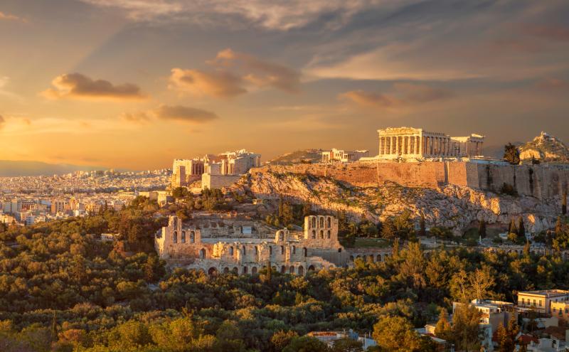 Panoramic view of the Acropolis of Athens, Greece, including the Parthenon and surrounding ancient ruins illuminated by warm evening sunlight, with the city of Athens stretching into the distance