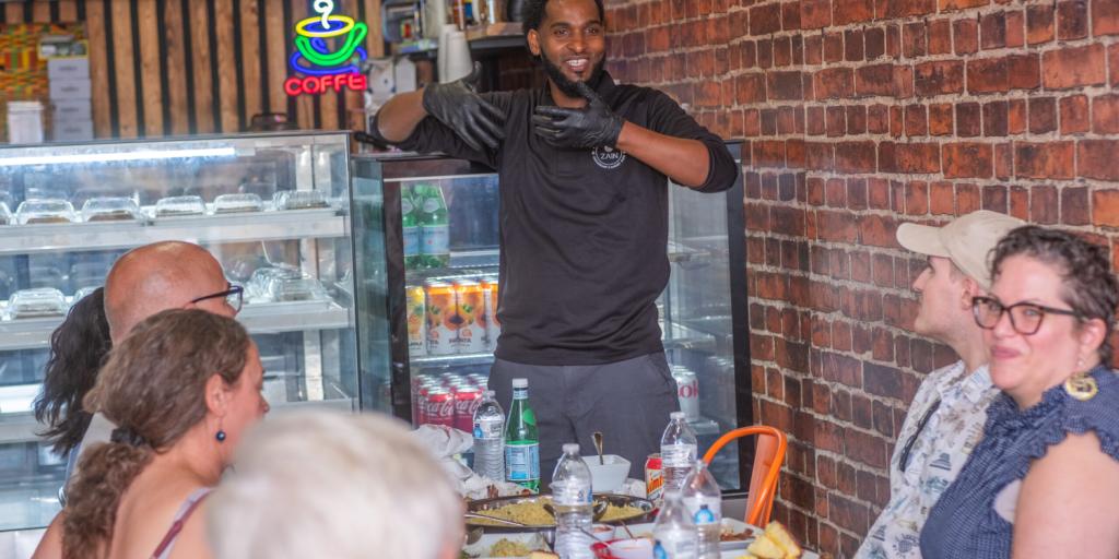 A guide at Culture Generation Food Tours in Kent speaks to a group of seated guests gathered around a table with plates of food inside a restaurant. The setting includes brick walls, a display case, and colorful menu signs.