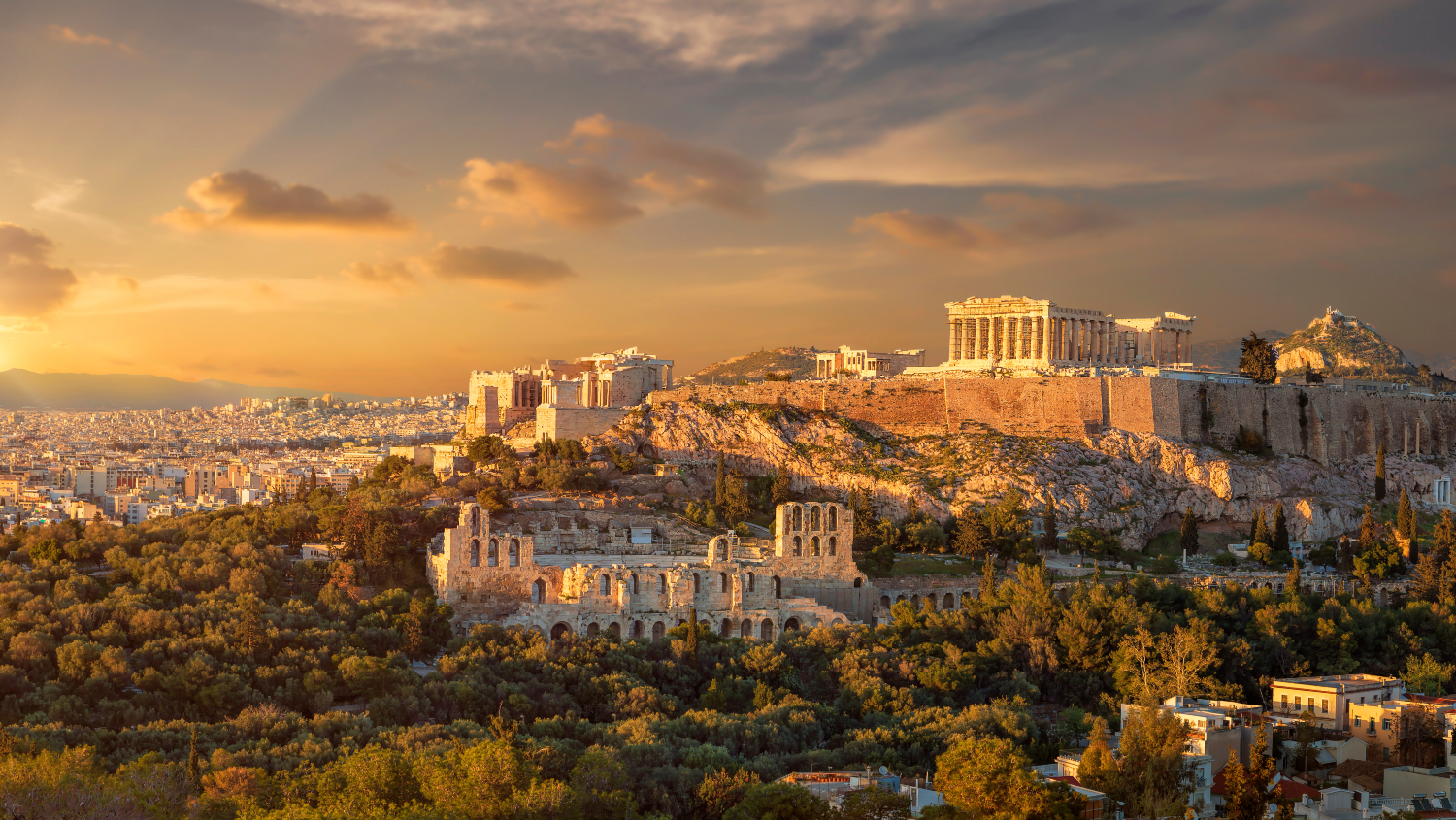 Panoramic view of the Acropolis of Athens, Greece, including the Parthenon and surrounding ancient ruins illuminated by warm evening sunlight, with the city of Athens stretching into the distance