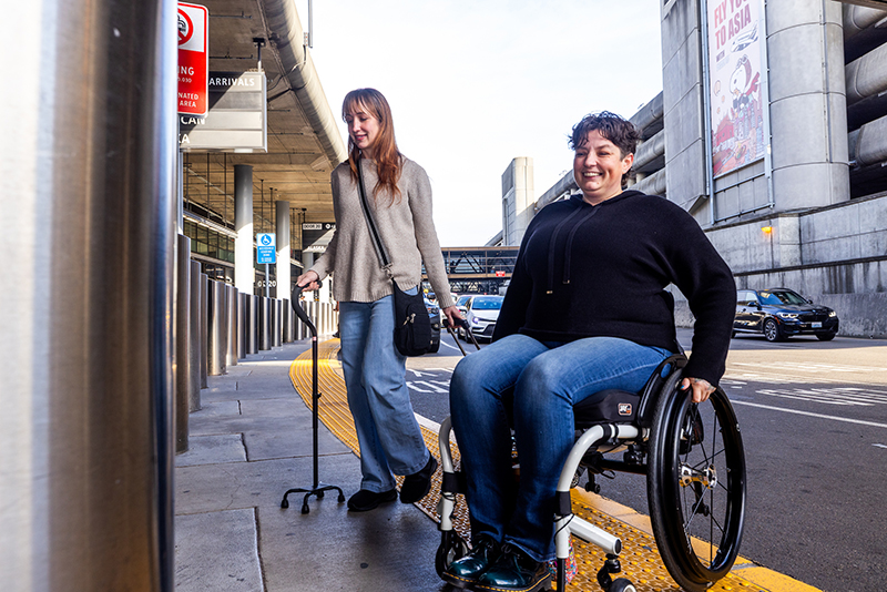 A traveler using a cane and a traveler using a wheel chair use the accessible loading zones at SEA.
