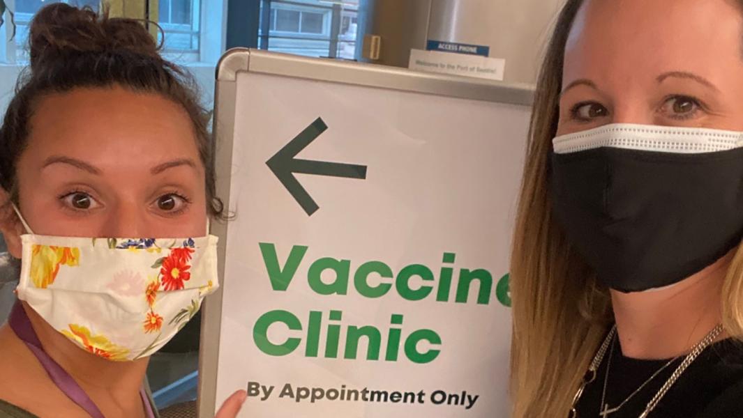 Two women wearing masks point to a sign pointing to a vaccine clinic at SEA Airport