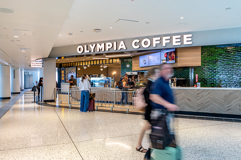 Image of the Olympia Coffee storefront at SEA with travelers walking by.