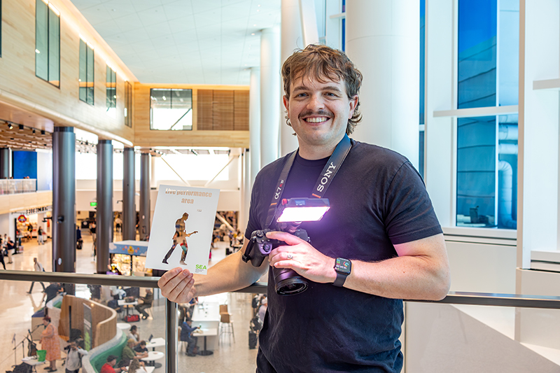 Rudy Willingham stands at Level 2 on the C Concourse Expansion holding a paper cutout of a magician and his camera.