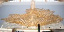 A large woven wooden sculpture shaped like a flaring canopy hangs high on an airport wall above gate signage.