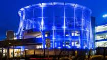 A circular parking structure wrapped in blue illuminated mesh is shown at night, with nearby road signs and foliage in the foreground.