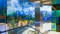 A colorful glass art installation with geometric patterns and nature‑inspired shapes surrounds an escalator in a brightly lit indoor space.