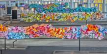 Colorful, abstract ribbon‑like sculptures stretch across an airport baggage claim area with conveyor belts and overhead signs in the background.