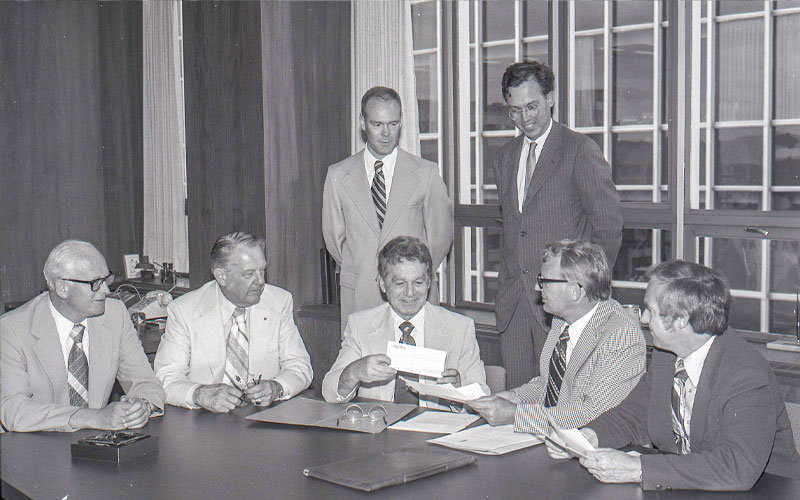 In a historic black and white photo, five men sit at a table the two in the middle holding a check and a deed. Two men look on behind them.