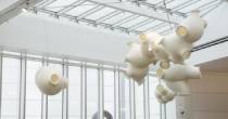 Suspended cluster of large white ceramic-like vessels hanging from the ceiling of a bright atrium with a glass roof and tall windows.