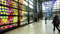 A large glass art installation featuring brightly colored circular patterns fills a tall window in a spacious indoor area, with another blue glass sculpture visible across the room as people walk through the space.