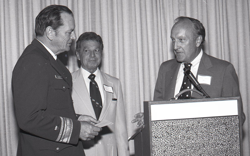 In a historic black and white photo three men stand at a podium talking.