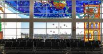 Large airport windows featuring colorful storybook‑style stained‑glass art above rows of empty seating.