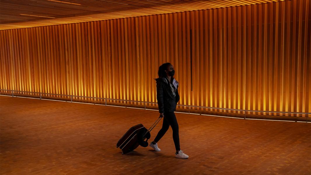 One passenger with rolling suitcase in tow walks through the warmly underlit wood paneling of the welcoming portal of the new IAF at Sea Airport