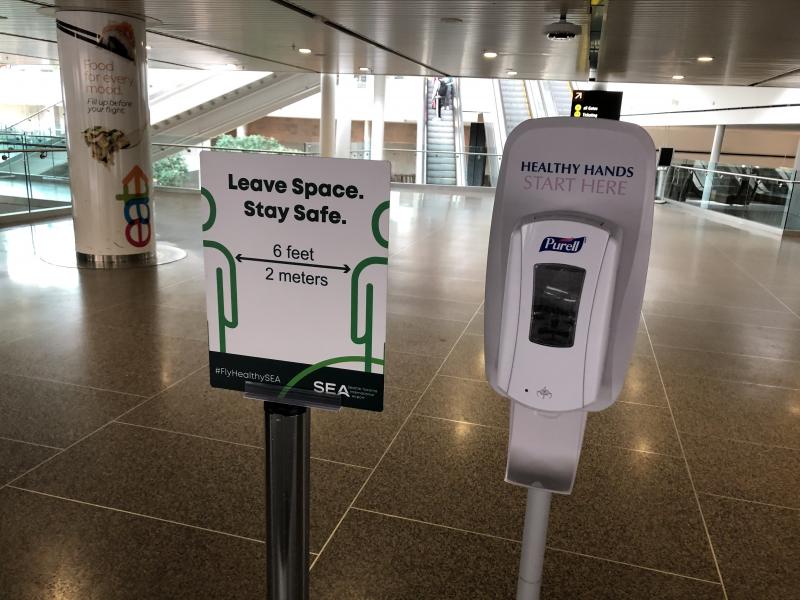 Hand sanitizer station next to a public information sign reminding passengers to sayt 6 feet apart, SEA Airport, May 2020