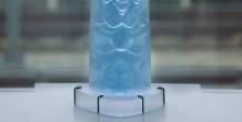 A light blue glass sculpture with carved abstract forms stands on a white pedestal in front of large windows.