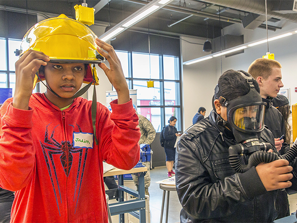Young man in orange pullover tries on a yellow protective helmet at a Youth Maritime Jobs event