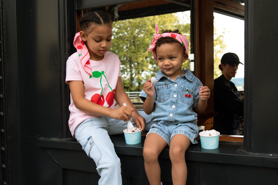 Two young girls sit side by side on a window ledge at an ice cream shop, each with a small cup of ice cream. The older girl wears a pink cherry-print t-shirt and the younger wears a denim romper with a pink headband, holding a spoon and looking at the camera. A blurred adult is visible through the window behind them.