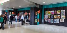 An airport hallway with gallery-style walls displaying colorful posters, with travelers walking past and others viewing the artwork.