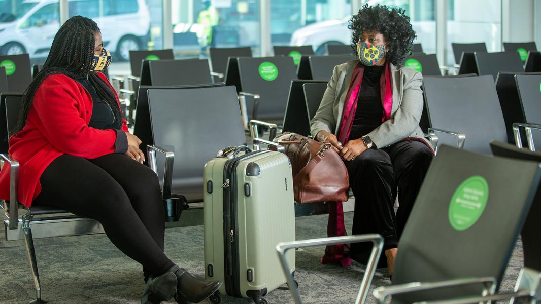 Two masked women in black with pops of red sit an empty seat apart to honor social distancing protocols at SEA Airport