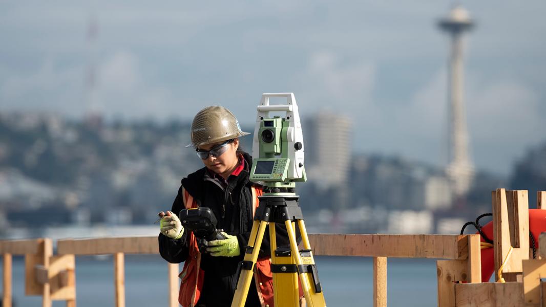 Surveyor in a yellow hard hat at Terminal 5 checks her instruments. Seattle skyline and Space Needle in the background