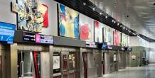 A transit station platform features a row of glass train doors beneath a series of large colorful abstract paintings mounted on the wall above.