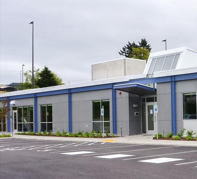 Exterior view of a single‑story gray building with blue trim, large windows, an accessible parking space, and landscaped plantings in front under an overcast sky.