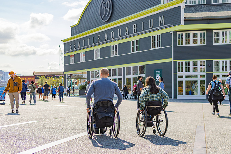 Two people using wheelchairs make their way along the waterfront in front of the Seattle Aquarium.