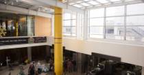 Airport security area with a tall yellow column beneath a large skylight and people moving through screening lanes.