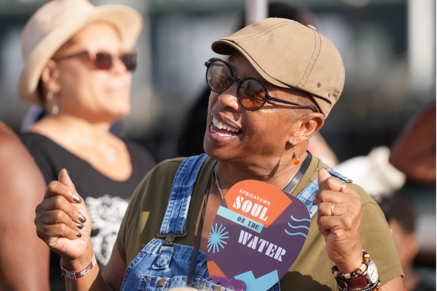 A joyful older woman wearing a tan baseball cap, round sunglasses, denim overalls, and orange drop earrings smiles broadly while holding an "Africatown Soul on the Water" event fan. A second woman in a hat and sunglasses stands blurred in the background.
