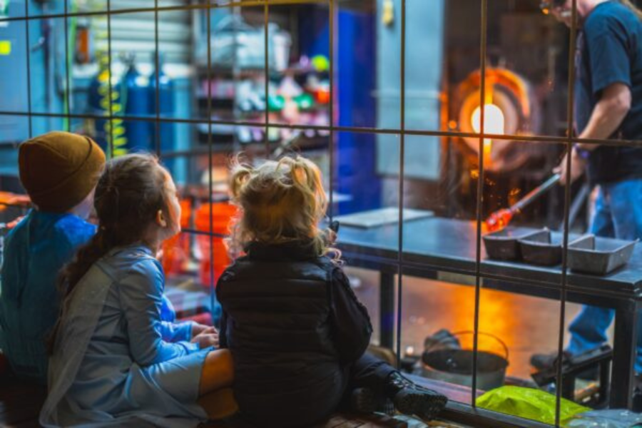 Three children sit side by side on a bench, viewed from behind, watching a glassblower work at a glowing furnace through a metal-grid safety barrier. The warm orange glow of molten glass illuminates the scene.