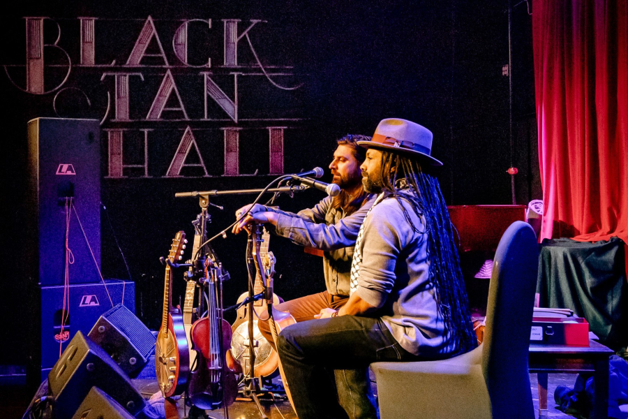 Two musicians sit on stage at Black Tan Hall, sharing a microphone. One has long hair and a beard; the other wears a wide-brimmed gray hat and has long dreadlocks. Several stringed instruments are visible on stands nearby.