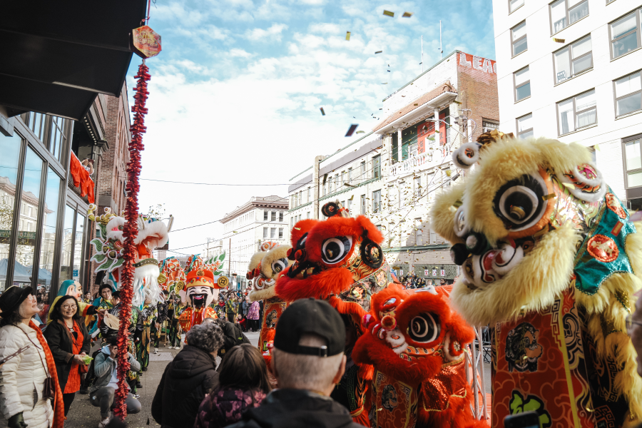 Vibrant Chinese lion dancers in red, gold, and teal costumes perform on a city street during a Lunar New Year celebration. A strand of red firecrackers hangs from a building, confetti fills the air, and a crowd of smiling onlookers lines the sidewalk.
