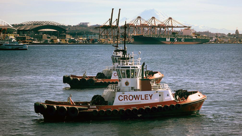 Two red and white tug boats in Puget Sound with Crowley on the side. The football stadium and port cranes are in the background.