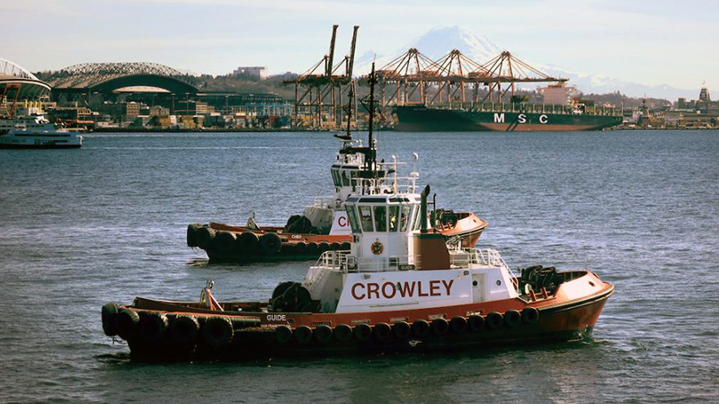 Two red and white tug boats in Puget Sound with Crowley on the side. The football stadium and port cranes are in the background.