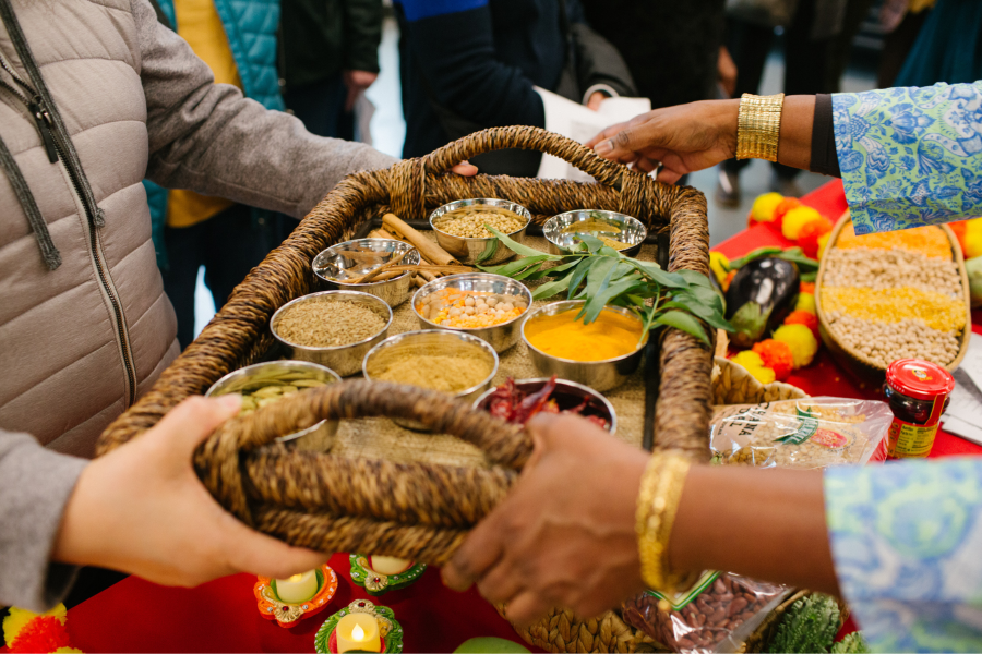 Two people pass a large woven basket tray holding small metal bowls filled with colorful spices, grains, and legumes. Marigold flowers, candles, and other cultural food items are visible on a red-draped table in the background.