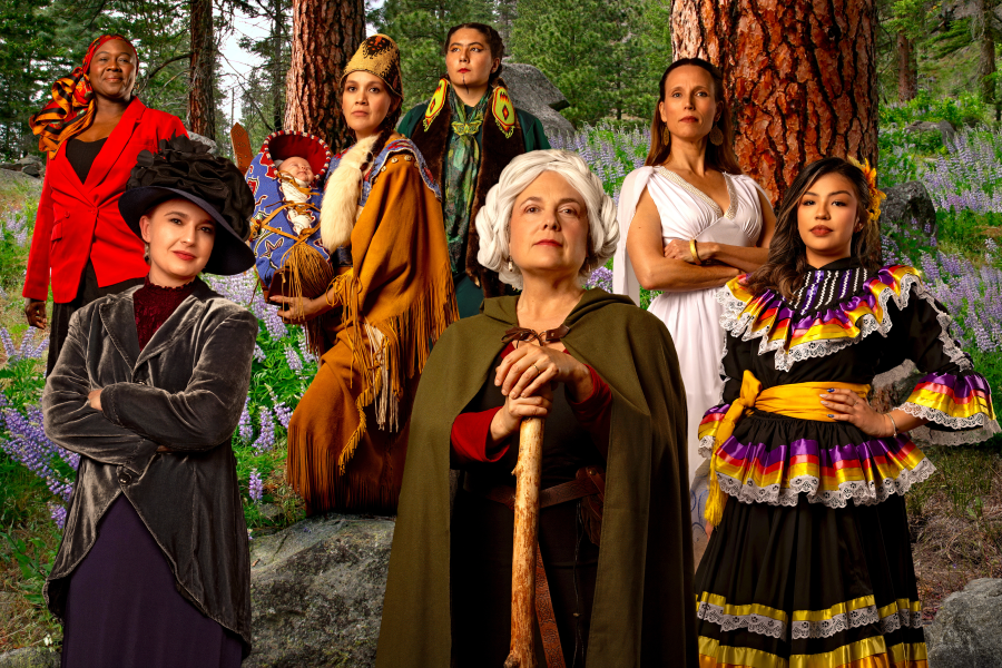 Eight women in diverse historical and cultural costumes pose together in a forest setting among tall pine trees and purple wildflowers. Costumes range from Native American regalia and a cradleboard to Victorian-era dress, a Greek-style gown, and a colorful folkloric skirt.