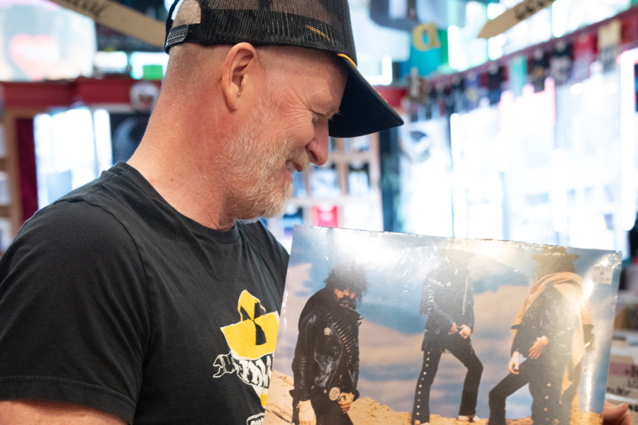 A middle-aged man with a short beard and trucker cap smiles as he examines a vinyl record album cover in a brightly lit music store. He wears a Wu-Tang Clan t-shirt, and shelves of records and merchandise are visible in the background.