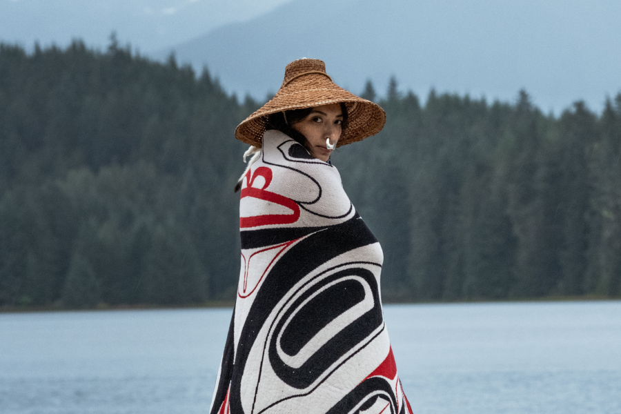 A person stands near a misty lake with forested mountains in the background, wrapped in a Northwest Coast Indigenous button blanket featuring bold black and red traditional formline designs. They wear a wide-brimmed woven cedar hat and look back over their shoulder toward the camera.