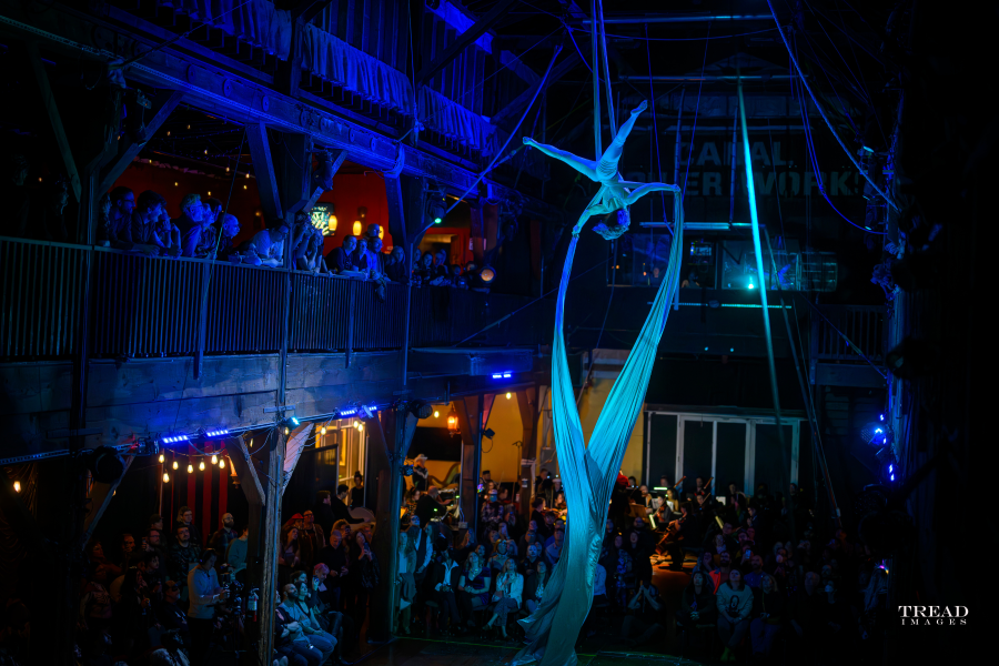 An aerial silk performer hangs dramatically upside down high above the crowd, wrapped in flowing blue fabric that cascades downward, lit by blue stage lighting. A large audience watches from the floor and an upper balcony of a rustic industrial venue.