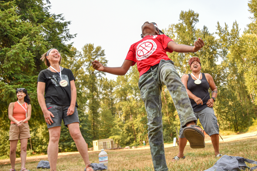 A youth in a red basketball t-shirt leaps exuberantly into the air with arms outstretched and head tilted back, celebrating outdoors. Two adults wearing event medals laugh joyfully nearby, with a third person smiling in the background. Tall evergreen trees frame the scene.