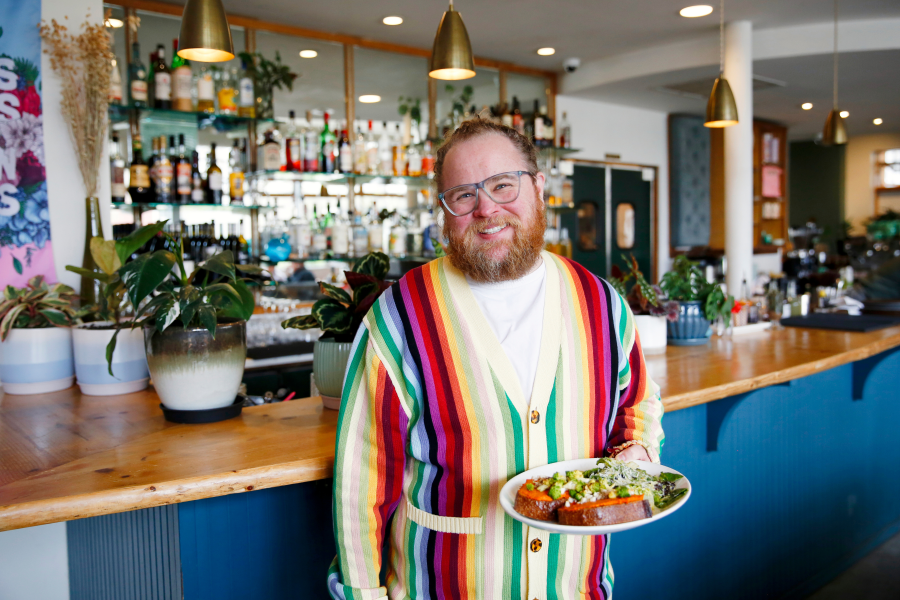 A smiling, red-bearded man wearing glasses and a bold multicolored striped cardigan holds a plate of food in a stylish restaurant. A full bar with shelves of spirits, hanging pendant lights, and potted plants is visible behind him.