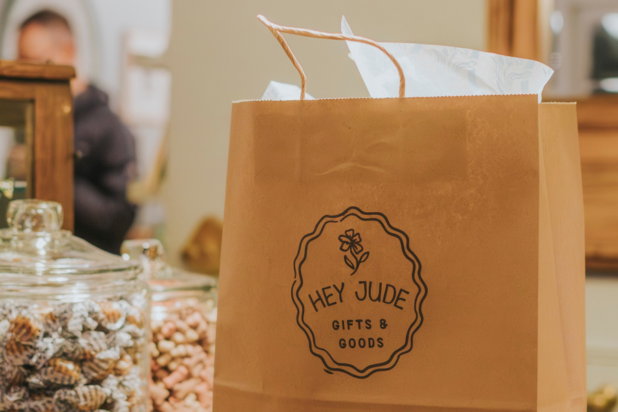 A close-up of a kraft paper shopping bag stamped with the logo "Hey Jude Gifts & Goods" featuring a simple flower illustration inside a decorative oval border. Glass jars filled with colorful candies and pasta are softly blurred in the foreground.