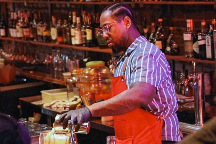 A focused bartender wearing glasses, a striped shirt, and a red apron pours a cocktail at a warmly lit bar. Shelves densely stocked with liquor bottles line the wall behind, and a large glass jar of infusing ingredients sits on the counter.