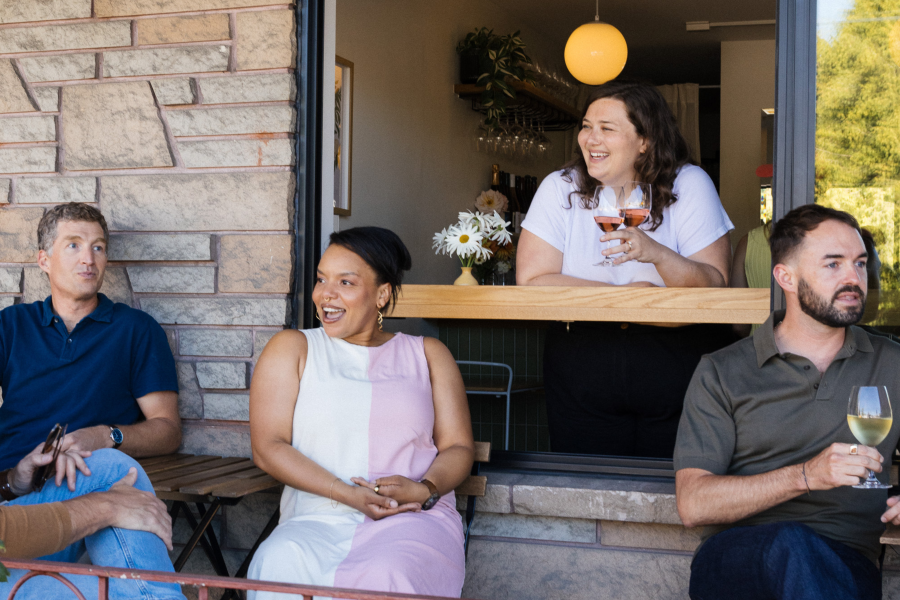 Four people socialize at what appears to be a small winery tasting room. A woman inside leans through a pass-through window holding two glasses of rosé, laughing with two guests seated outside on steps and another seated on a bench. A small vase of daisies sits on the ledge.