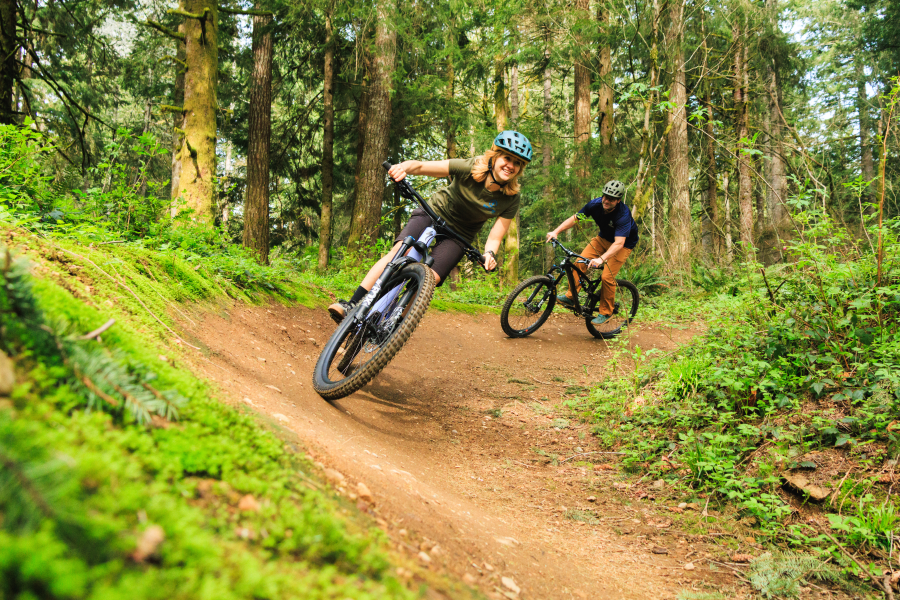 Two mountain bikers wearing helmets navigate a banked dirt trail through a lush, moss-covered Pacific Northwest forest. The lead rider, a woman in a teal helmet, grins at the camera as she carves through the turn, with a second rider close behind.