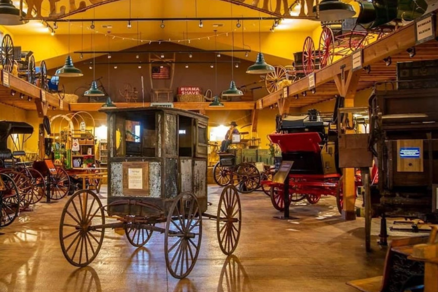 The interior of a carriage museum with a warm golden color scheme, displaying multiple antique horse-drawn vehicles on a polished wood floor. A weathered wooden stagecoach occupies the foreground, a bright red open carriage sits to the right, and additional wagons and wagon wheels are displayed on an upper mezzanine level. Vintage signs including "Burma Shave" hang from the ceiling alongside green pendant lights.
