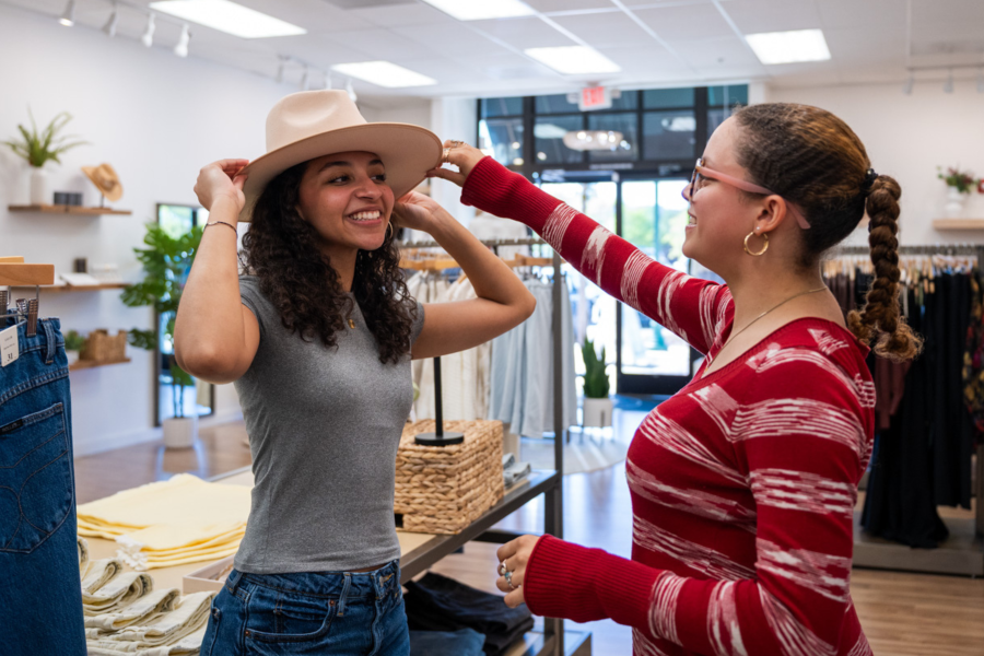 Two young women laugh together in a bright, modern clothing boutique as one tries on a wide-brimmed tan felt hat. The friend in a red striped sweater reaches over to adjust the brim. Racks of clothing, folded jeans, and woven baskets are visible in the cheerful, plant-accented store behind them.